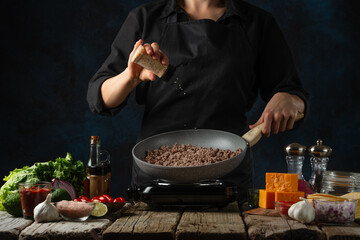 Wall Mural - Close-up view of professional chef in black uniform salts meat filling for cooking tacos on dark blue background. Traditional mexican cuisine. Concept of tasty street food. Frozen motion.