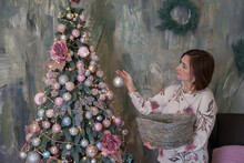 A Woman Near Christmas Tree Holding A Box With Christmas Balls. A Pretty Female With Brown Hair In Sweater Decorating The Christmas Tree With Ball. Pink Christmas Decor