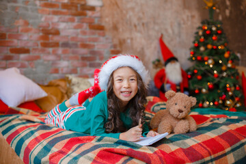 Wall Mural - little Asian girl in pajamas near the Christmas tree writes a letter to Santa Claus Christmas, new year