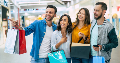 Wall Mural - Happy group of excited beautiful modern stylish friends in casual wear with paper bags are walking in the mall during shopping.