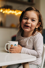 Wall Mural - Caucasian toddler girl drinking cocoa from mug at home. Christmas bokeh lights