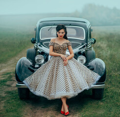 Young beautiful sexy woman in pin-up style clothes posing near black retro car. Polka dot white dress, vintage hairstyle, red high heels. Background road green nature fog. Girl fashion model driver