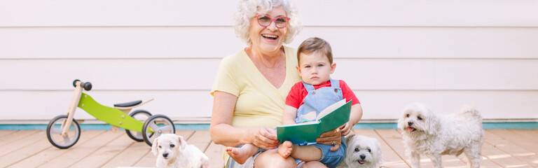 Grandmother reading book to grandson boy at home backyard. Bonding of relatives and generation communication. Old woman with baby spending time together outdoors. Web banner header.