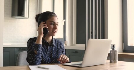 Canvas Print - Happy indian ethnicity businesswoman working on laptop at home office, consulting client distantly by smartphone call, helping with problem solution or discussing working issues with colleagues.