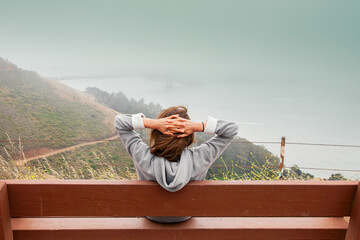 Back view of a girl in a gray hoodie. Woman sitting on a bench and admires the panorama of the city and the bridge (Golden Bridge in San Francisco) from a high mountain. Vacation and travel concept