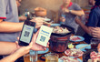 © Koonsiri - Hand holding smartphone to scan Qr code payment tag with blurry grilled pork and meat on the stove, Thai food buffet payment tag. QR code payment concept to many friends at a party in a  restaurant.
