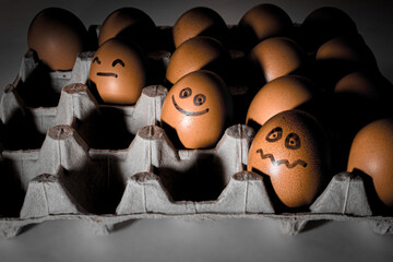 Many orange spotted brown chicken eggs in carton open box container in light with dark shadows. Three eggs with black drawn smile, emotion faces