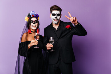 Wall Mural - Joyful boyfriend and girlfriend drink champagne and celebrate Halloween in image of Mexican-style bride and groom