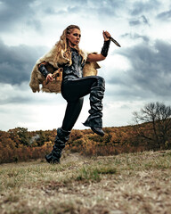 A young girl with long blond hair in a historical Viking costume with a weapon in nature under a threatening autumn sky.