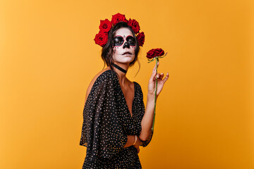 Wall Mural - Charming female model in elegant dress enjoying scent of roses. Portrait of girl with Halloween makeup posing in orange room