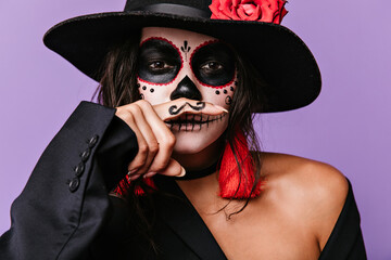 Wall Mural - Mischievous brown-eyed girl with face art is having fun by putting painted mustaches to her mouth. Photo of stylish woman in black outfit with red details