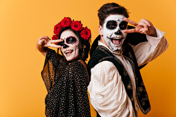 Wall Mural - Cheerful young man and woman are having fun on orange background and show peace sign. Portrait of painted couple in national Mexican costumes