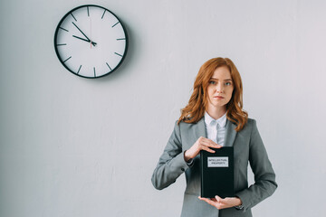 Redhead female lawyer holding book with intellectual property lettering, while standing near wall clock on grey, stock image