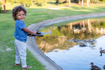 Cute little boy smiling as he learns to fish in a pond at an outdoor park. Young black boy holding a fishing pole as he attempts to catch a fish