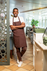 Poster - Cheerful young waiter in workwear leaning against armchair by served table