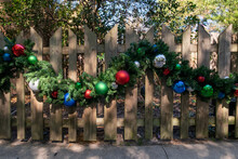 A Light Coloured Textured Wooden Paling Fence With A Christmas Garland Looped Along The Middle Of The Barrier. The Garland Is Made Of Evergreen Branches And Has Colourful Christmas Balls Intertwined.