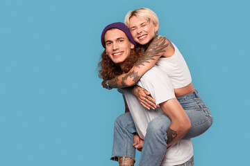 Freestyle. Young tattooed couple standing isolated on blue girlfriend riding boyfriend's back hugging smiling happy