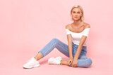 Fototapeta Panele - Freestyle. Young blonde woman in jeans and top sitting isolated on pink posing to camera pensive