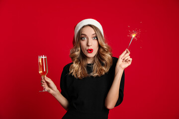 Sticker - Surprised young woman wearing Santa hat with glass of champagne and sparkler on red background. Christmas celebration