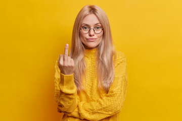 Wall Mural - Serious self confident blonde woman shows fuck off sign or middle finger gesture wears round spectacles and long sleeved jumper being impolite and rude poses against yellow studio background