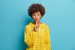 Surprised dark skinned young woman makes shush gesture asks to keep information in secret shows hush sign has shocked face expression wears casual yellow sweater isolated over blue background.