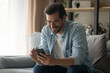 © fizkes - Smiling young Caucasian man sit on couch in living room look at smartphone screen texting or messaging on gadget online. Happy male relax at home browse wireless internet on cellphone device.