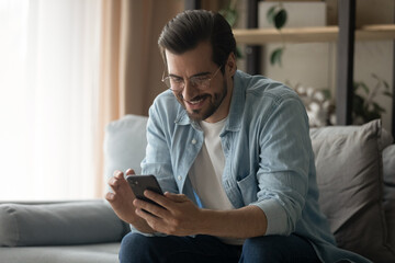 Wall Mural - smiling young caucasian man sit on couch in living room look at smartphone screen texting or messagi