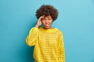 Wall Mural - Displeased curly haired Afro American woman keeps hand on temple suffers migraine or severe headache frowns face and has problems wears casual knitted yellow jumper isolated over blue background