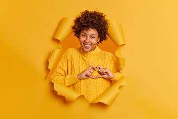 Wall Mural - Lovely smiling woman shapes heart gesture being in good mood expresses love to you says be my valentine confesses in truthful feelings wears yellow sweater poses through paper background in hole
