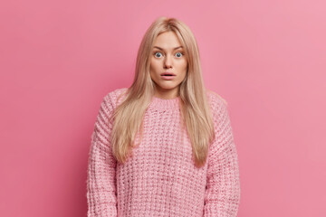 Wall Mural - Shocked blonde young European woman has stupefied face expression stares bugged eyes cannot believe in shocking horrible news wears knitted sweater poses against pink background. Omg concept