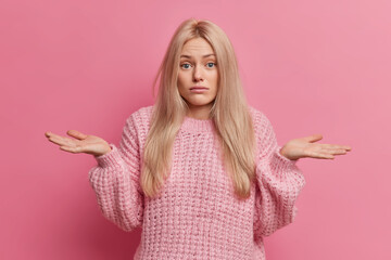 Wall Mural - Uncertain blonde woman spreads palms and stands doubtful indoor cannot make choice between two options wears knitted warm sweater isolated over pink background. Doubtful female model shrugs shoulders