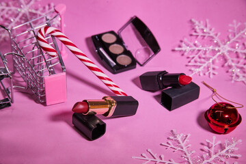 Wall Mural - Creative concept with shopping trolley with makeup on a pink background.  Red lipstick, eyeshadows. 
With a candy cane, red Christmas ball and snowflakes. Concept of Christmas shopping for beauty.