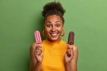 Wall Mural - Photo of cheerful dark skinned African American woman holds strawberry and chocolate ice cream smiles broadly dressed in casual yellow t shirt poses against green background. People summer concept