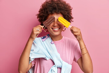 Wall Mural - Horizontal shot of happy curly haired woman has fun during summer day covers eyes with delicious ice cream and smiles broadly wears t shirt and sweater tied over shoulder poses indoor over pink wall