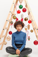 Vertical Image Of Shocked Curly Haired Woman Sits Crossed Legs And Stares Startled At Camera Stunned New Year Is Already Tomorrow Has Too Much To Do Sits Near Ladder With Toy For House Decorating