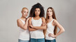 Three confident young diverse women wearing white shirts looking at camera while posing together isolated over grey background