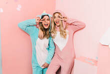 Winsome Girls In Kigurumi Standing On Pink Background. Gorgeous Sisters In Pajamas Posing With Peace Sign.