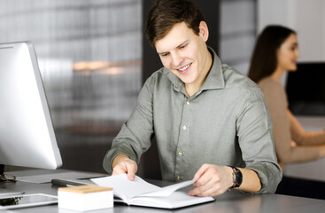 Smiling young businessman and programmer in a green shirt is flipping his notebook, while sitting at the desk in a modern cabinet together with a female colleague at the background. Concept of
