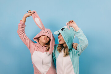 Laughing attractive ladies posing in kigurumi. Carefree girl in bunny costume standing on blue background.