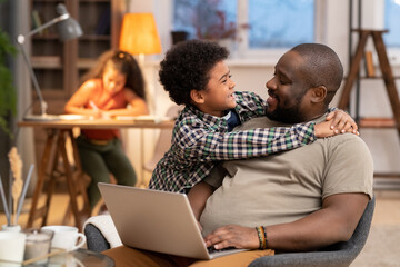 Canvas Print - Happy affectionate African boy embracing his father with laptop against daughter