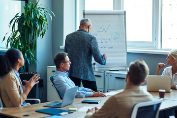 Wall Mural - Mature businessman writing something on flipchart while having a meeting with multiracial colleagues in the modern office