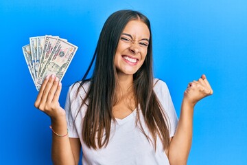 Young hispanic woman holding dollars screaming proud, celebrating victory and success very excited with raised arm