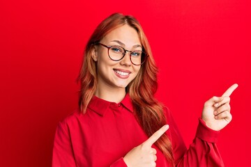 Young beautiful redhead woman wearing casual clothes and glasses over red background smiling and looking at the camera pointing with two hands and fingers to the side.