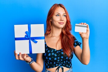 Sticker - Young redhead woman holding gift and credit card smiling looking to the side and staring away thinking.