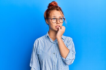 Canvas Print - Young beautiful redhead woman wearing casual clothes and glasses over blue background looking stressed and nervous with hands on mouth biting nails. anxiety problem.