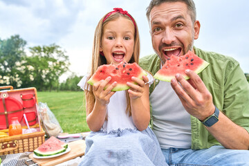 Portrait of happy dad and his adorable little daughter looking at camera while eating watermelon, family having a picnic in the green park on a summer day