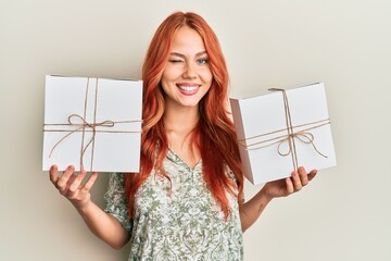 Young redhead woman holding gifts winking looking at the camera with sexy expression, cheerful and happy face.