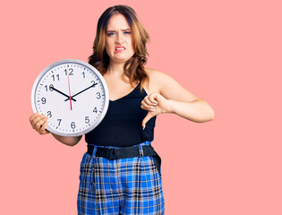 Poster - Young beautiful caucasian woman holding big clock with angry face, negative sign showing dislike with thumbs down, rejection concept