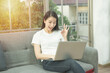 © mkitina4 - Freelance Jobs workation concept.Positive smile asian girl sitting on sofa with laptop and showing ok sign.