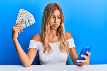 Sticker - Beautiful blonde young woman holding dollars and using smarpthone depressed and worry for distress, crying angry and afraid. sad expression.